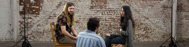 Nick sits in front of a man and woman sitting across from each other on wooden chairs with intrigued expressions.