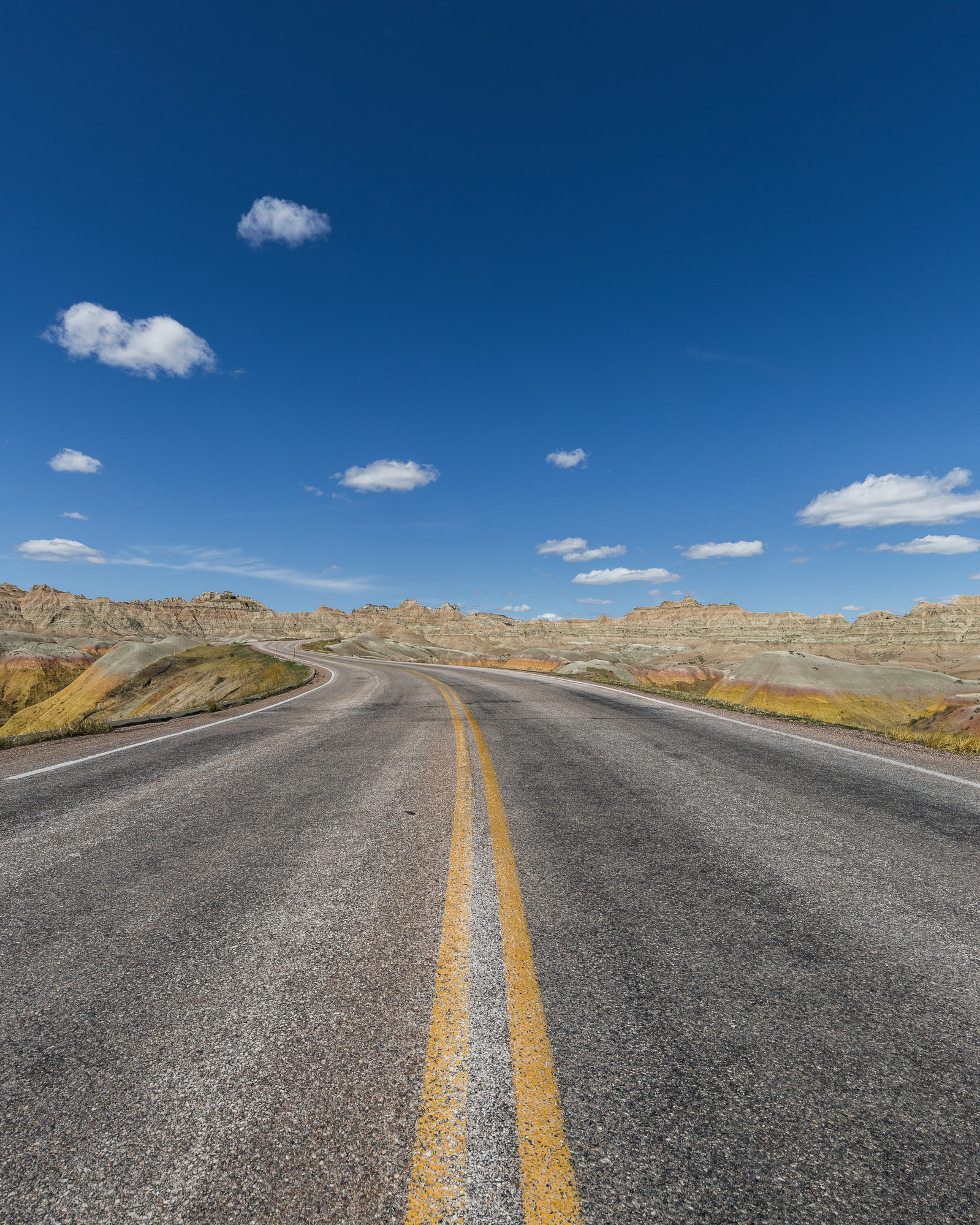 Low-angle view of a paved road winding through the colorful rock formations of Badlands National Park, South Dakota, under a clear blue sky with scattered clouds.