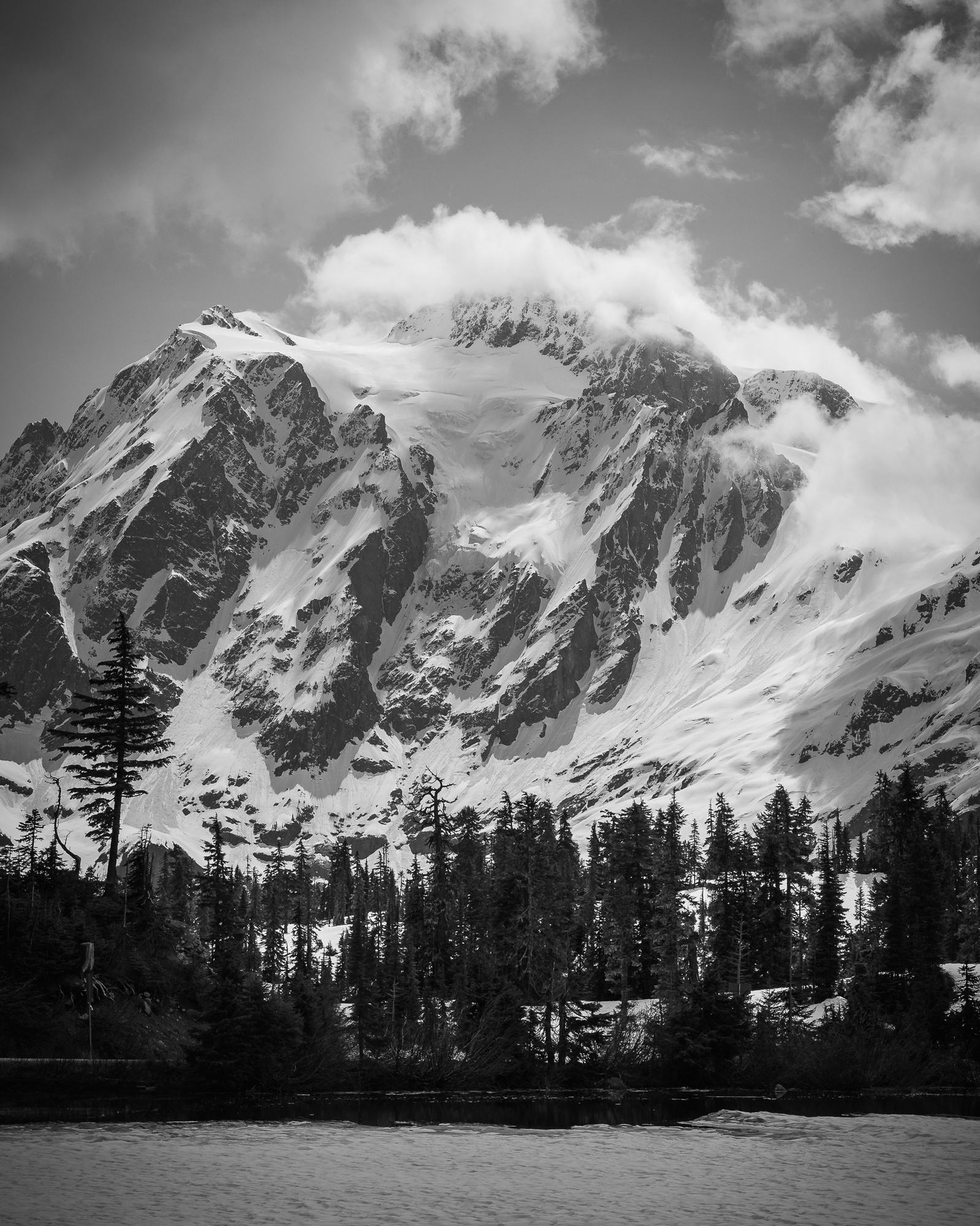 Snow-covered Mt. Shuksan rises behind a forest of evergreens, with low clouds brushing its rugged peak and a still lake in the foreground.