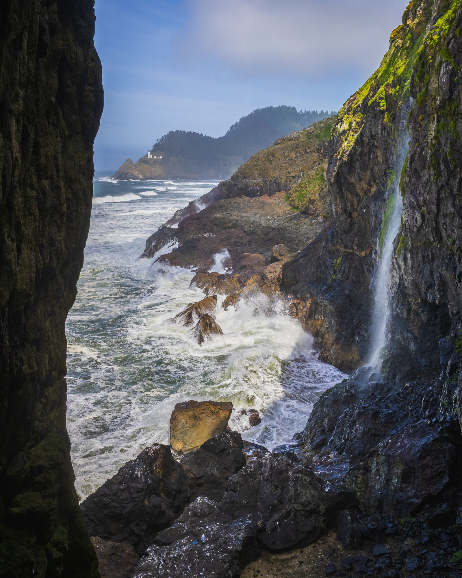 View of the Oregon coast with waves crashing against rocky cliffs, a small waterfall spilling down the right side, and Heceta Head Lighthouse visible in the distance.