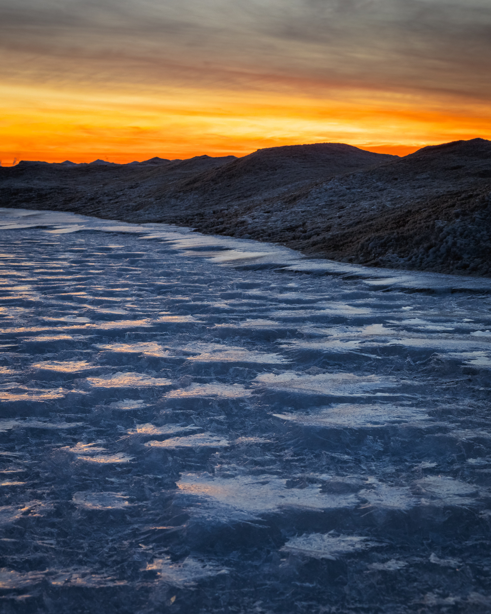 Frozen shoreline of Lake Michigan at sunset, with textured ice in the foreground and snow-covered dunes silhouetted against an orange and yellow sky.