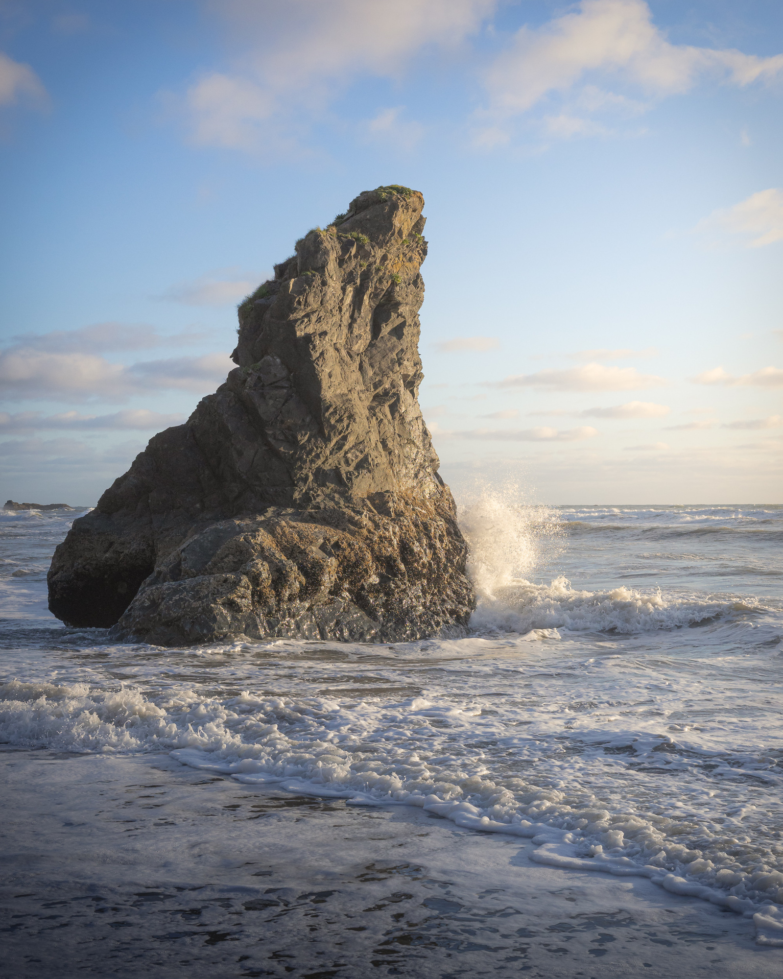 Sea stack at Olympic National Park with waves crashing against its base during golden hour, under a partly cloudy sky.