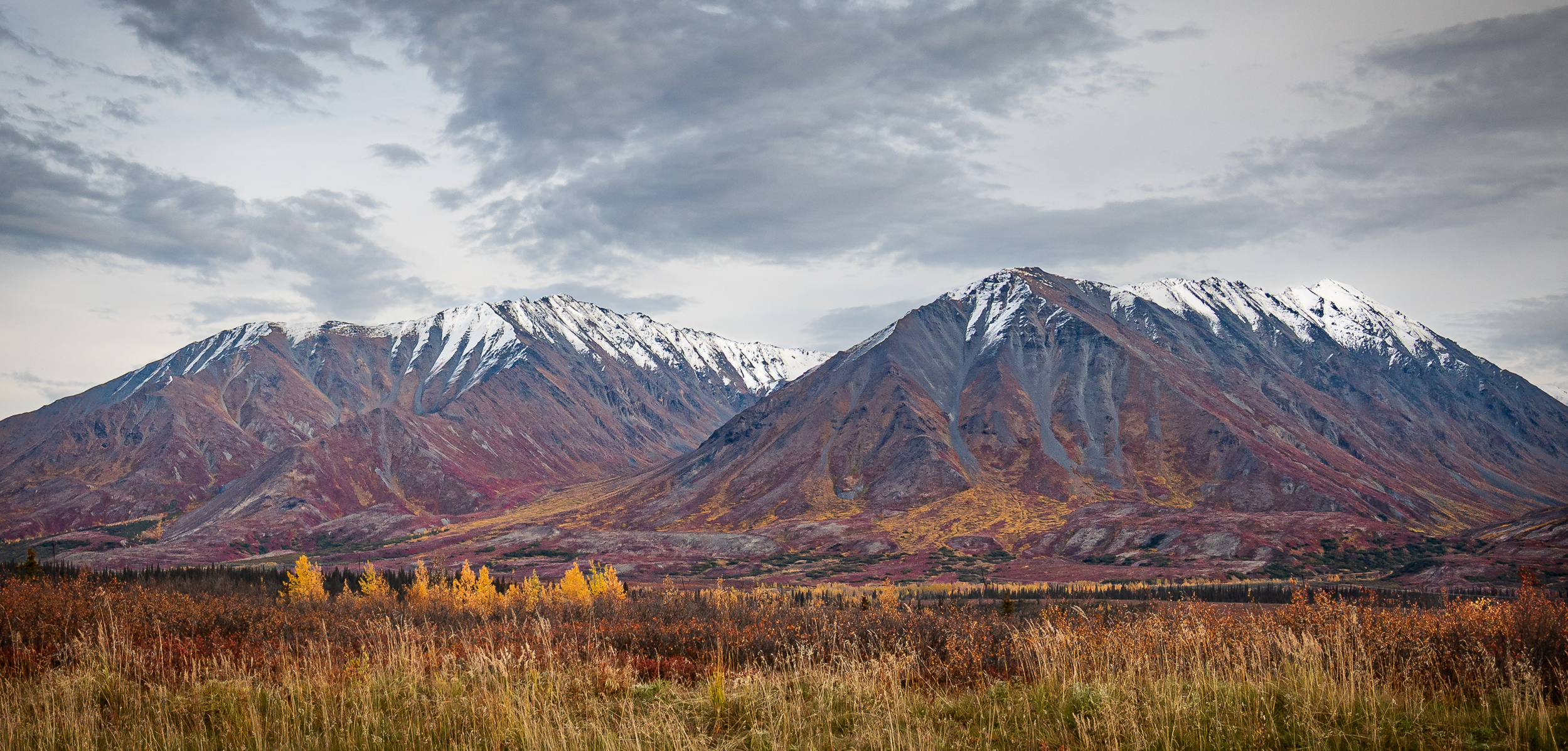 Two rugged, snow-dusted mountains rise above a valley of autumn tundra in Alaska, with streaks of red, orange, and gold vegetation beneath a cloudy sky.