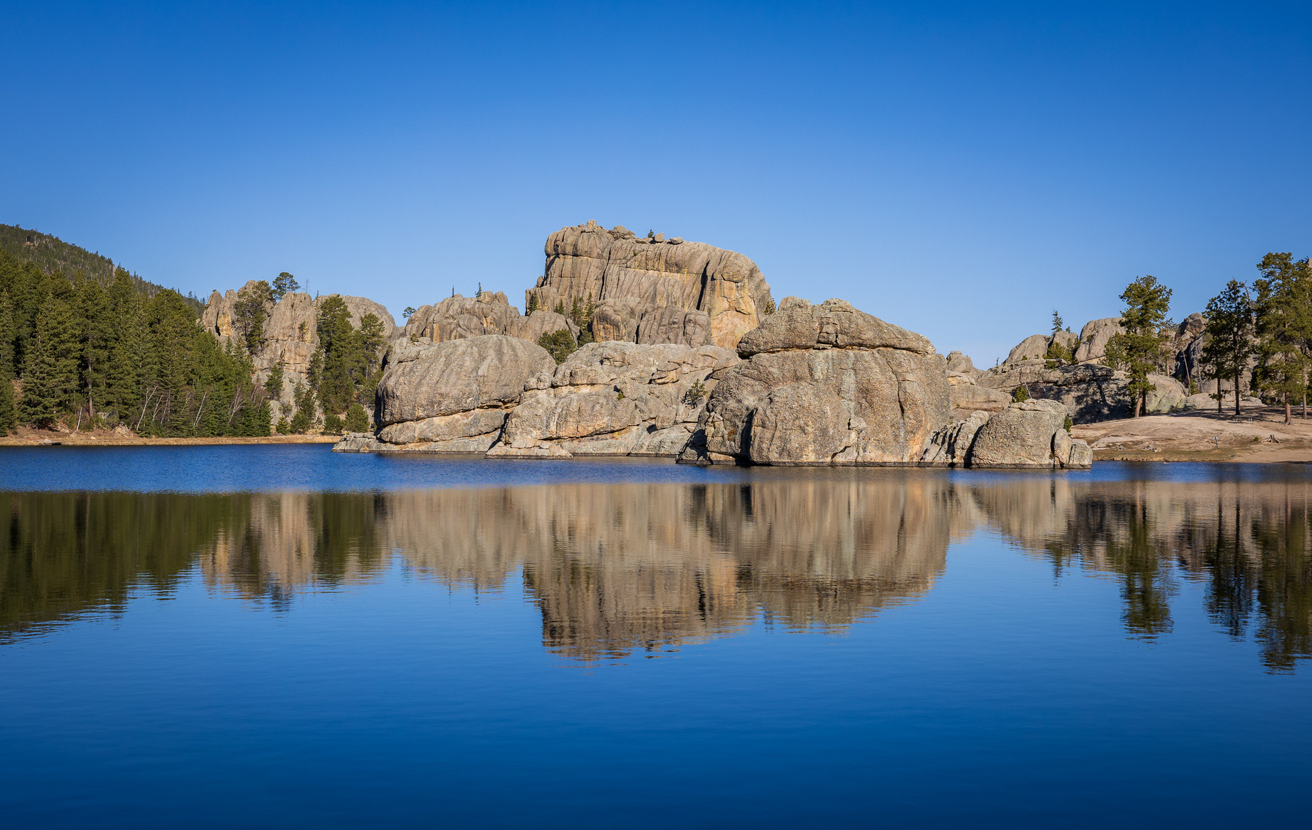 Granite rock formations reflected in the calm blue water of Sylvan Lake in Custer State Park, South Dakota, under a clear sky.