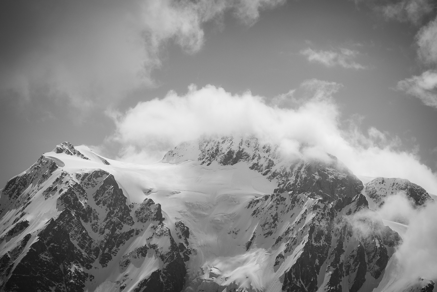 Black and white photo of a snow-covered mountain peak partially obscured by clouds, with rugged ridgelines and glacier formations.