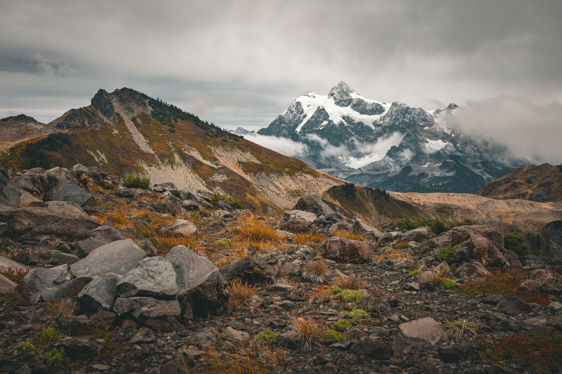 View of Mount Shuksan in Washington, with autumn alpine vegetation and rocky terrain in the foreground under overcast skies.