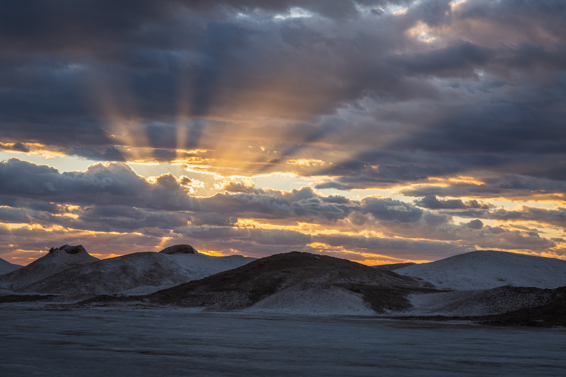 Winter sunset over frozen dunes along Lake Michigan, with sunbeams breaking through heavy clouds and illuminating the icy landscape.