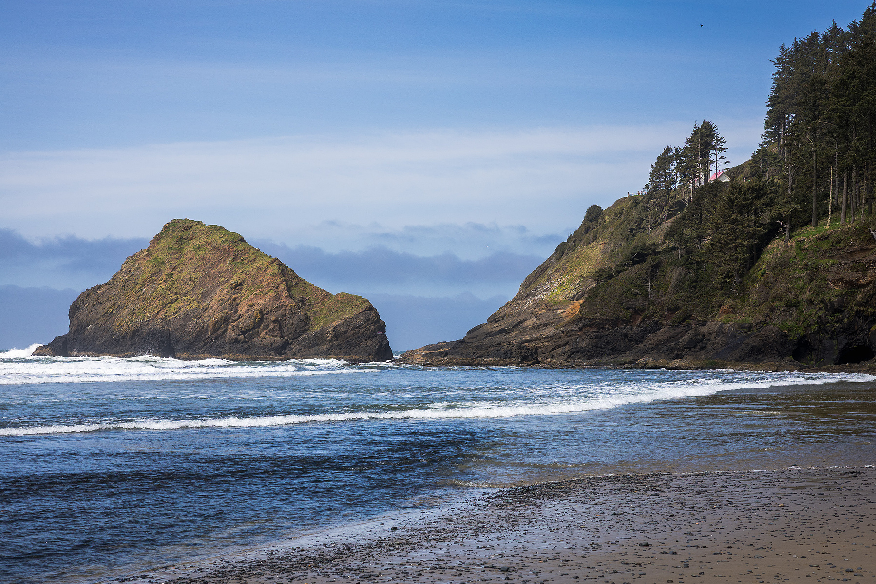 Coastal landscape photo from the Oregon Coast featuring a sea stack, ocean waves, and forested cliffs under blue skies. Nature photography highlighting Pacific Northwest scenery.