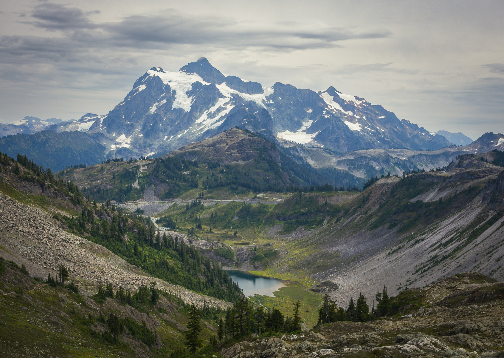 View of Mount Shuksan in Washington with snow-covered peaks, a glacial valley, and a small alpine lake below under a cloudy sky.