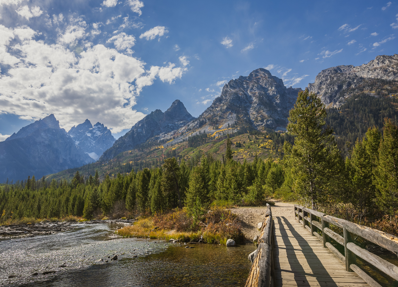 Jenny Lake footbridge in Grand Teton National Park crossing a creek with forested slopes and the Teton Range in the background under a partly cloudy sky.