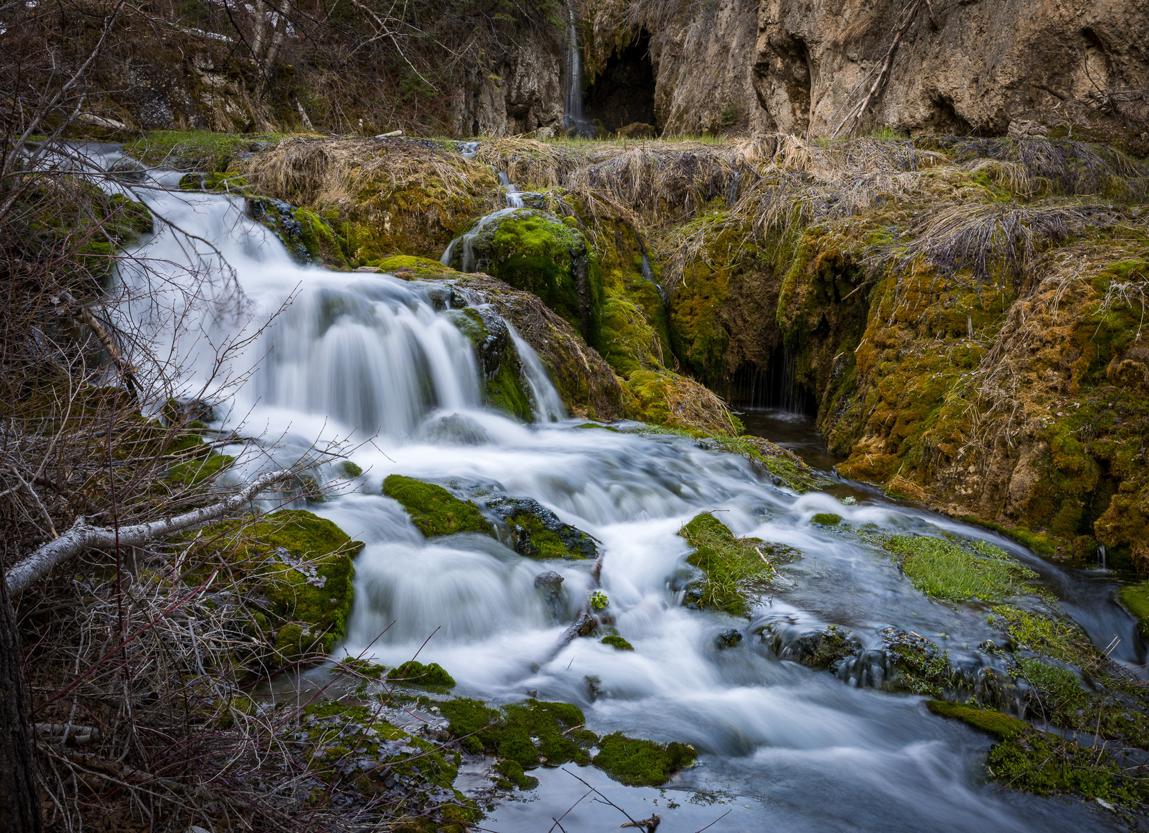 Roughlock Falls in South Dakota, with cascading water flowing over moss-covered rocks and grassy terraces surrounded by cliffs and forest.