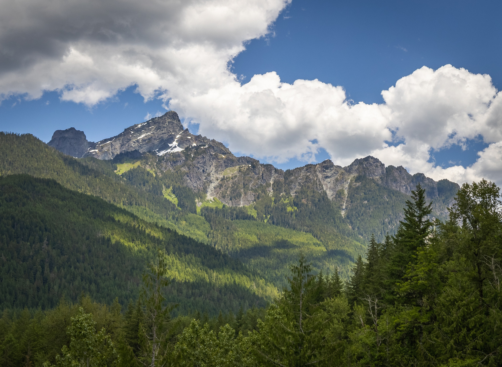 Mountain peaks in the Cascade Range rising above a dense evergreen forest under partly cloudy skies in Washington.