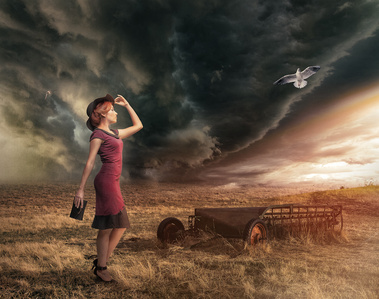 An environmental portrait of a young woman in a red dress holding the brim of her dark hat looking off into the distance as a prairie storm brews in the distance