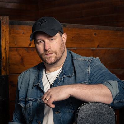 A male in a denim jacket on a white t-shirt wearing a cap and resting his arm on a guitar case.