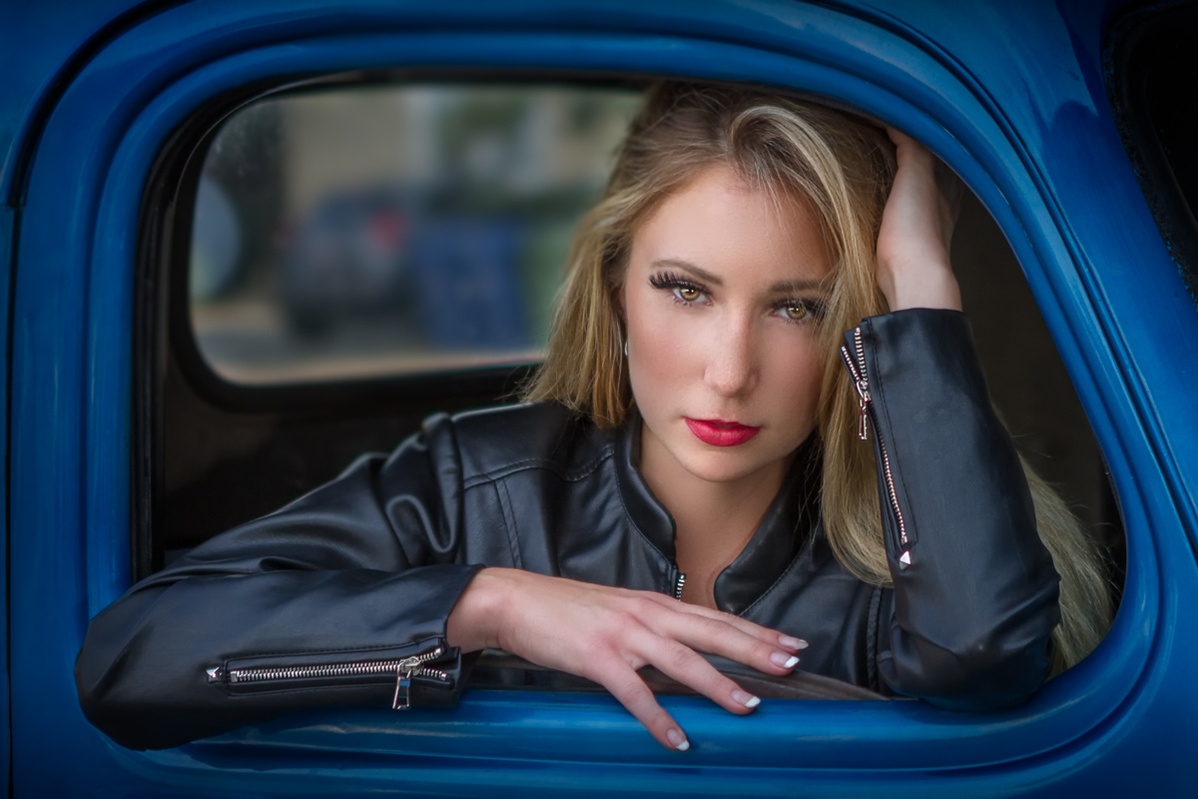 An attractive blond woman wearing a denim jacket looking out of the window of a blue truck