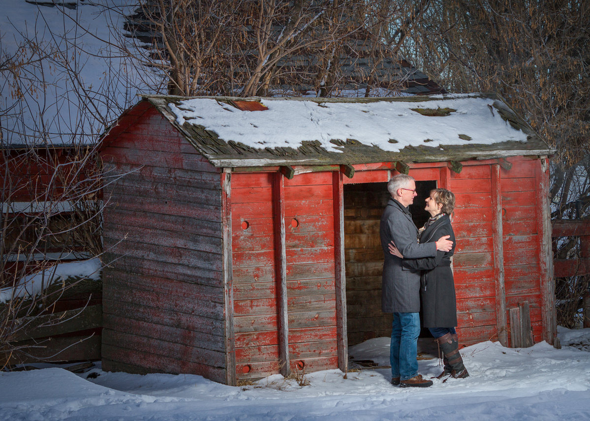 A farm yard winter photo of a man and wife embracing in front of a red shed covered with some snow