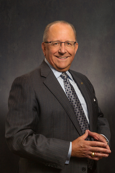A business-style headshot of a gentleman with glasses wearing a dark grey suite and light blue shirt with dark ties all on a grey background.