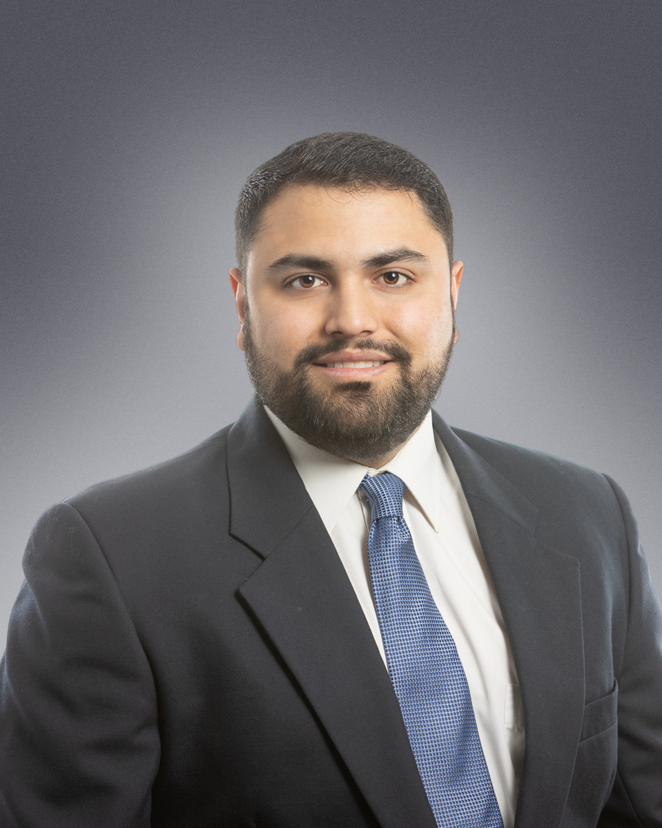 Headshot of a man wearing dark suite with a white shirt and blue tie on a grey background