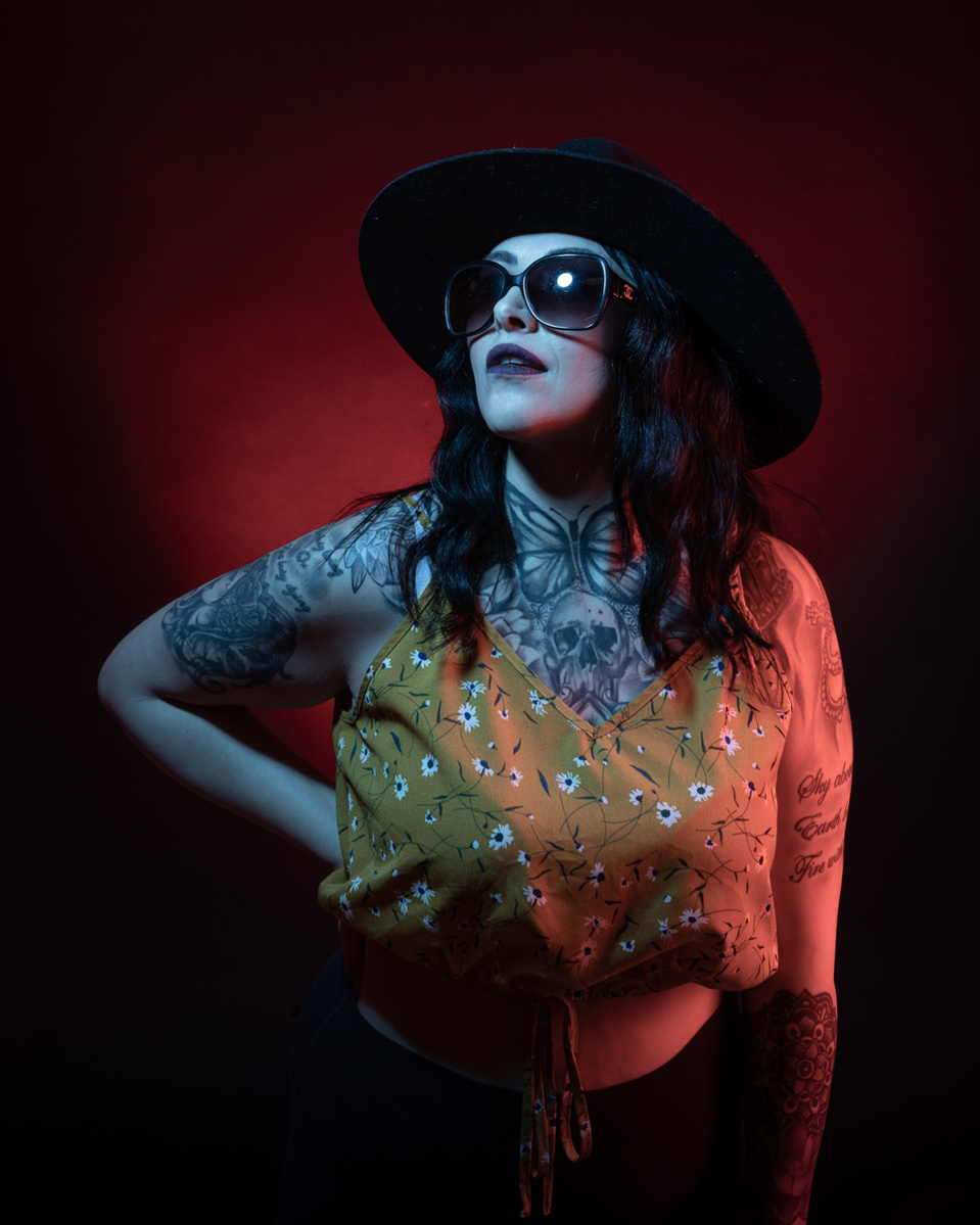 A portrait of a woman wearing a black hat and dark sunglasses on a dark red background