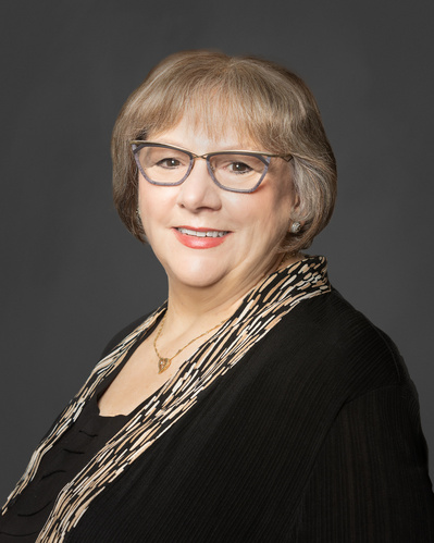 Portrait headshot of an older woman wearing glasses and a dark top against a grey background.