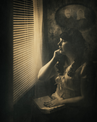 A pictorialism-styled monochromatic photo of a woman in victorian era clothing contemplatively looking out a shaded window.