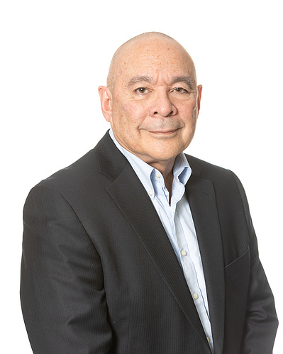 A headshot portrait of an older bald gentleman wearing a dark suit jacket over a light blue open-collar shirt all on a white background.