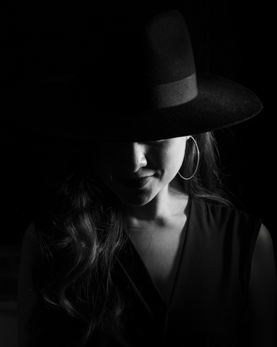 A low-key black and white headshot of a young woman wearing a black hat and looking downwards.