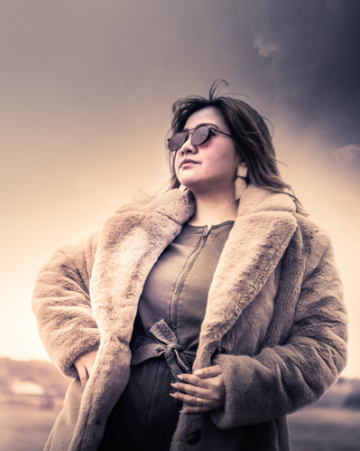 An environmental duo-tone portrait of a dark-haired woman with sunglasses wearing an open fur jacket.