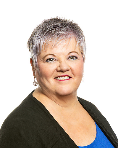 A headshot portrait of a woman with short great hair wearing a black sweater over a blue top against a white background.