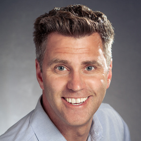 An headshot of an actor smiling and wearing a blue shirt on a gradient grey background