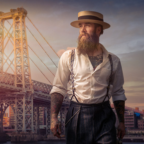 A warm sunset creative portraiture shot of a gentleman wearing vintage clothing and a straw hat looking off into the distance with a large city bridge in the background
