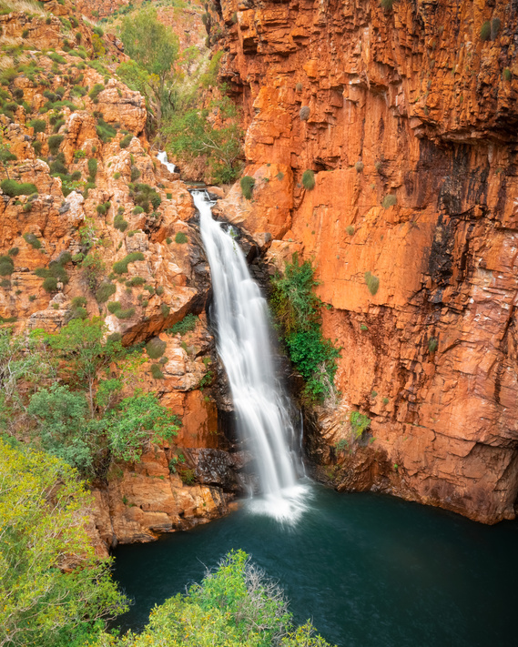 A large waterfall cascades off of the orange sandstone cliff into the freshwater pool below.