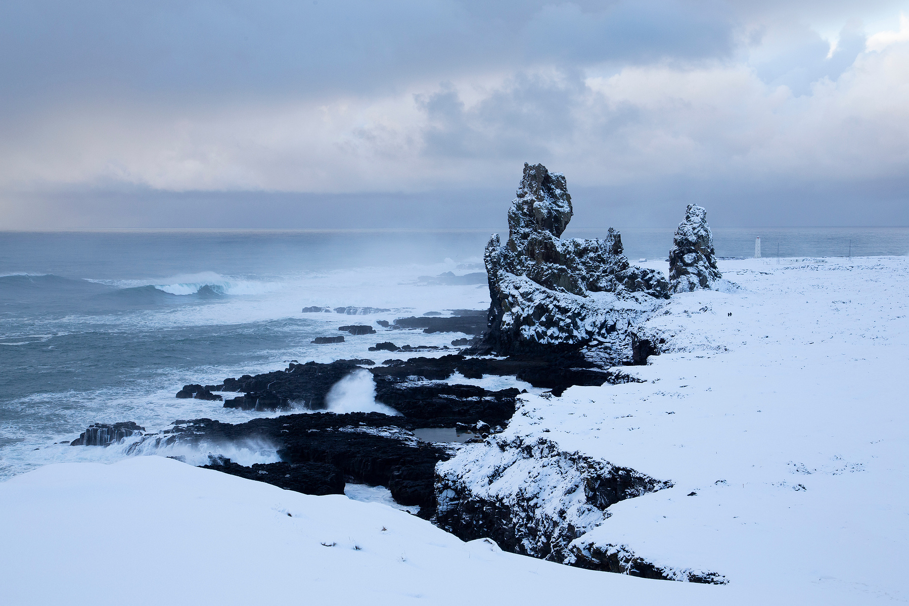 The Londrangar Cliffs on the Snaefellsnes peninsula in Iceland just after a snow storm. The snow highlights the jagged shape of the black volcanic rock towers while North Atlantic Ocean waves crash upon the shore.