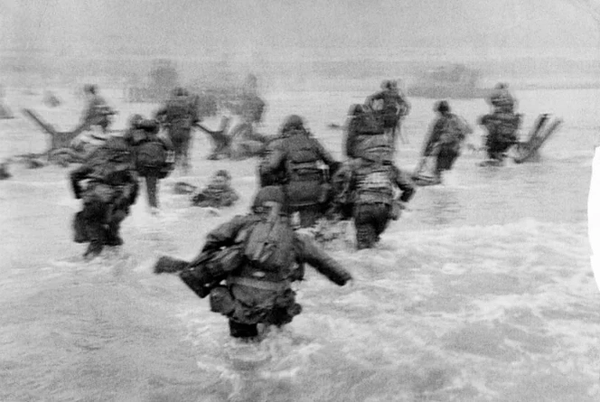 La memorable imagen de Robert Capa durante el Desembarco de Normandía muestra valientes soldados avanzando a través del agua bajo fuego enemigo, simbolizando la audacia y sacrificio en la lucha por la libertad.