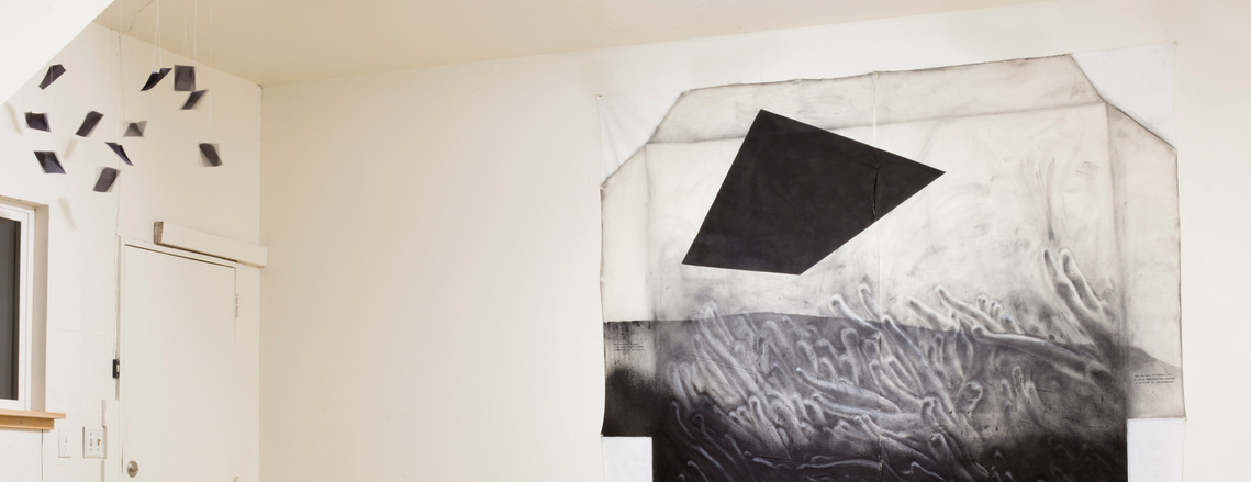 8 small swaying black paper kites hang from the ceiling in the left corner of a white room. To the right, a large drawing  is half of the image. The drawing contains a black four sided shape above a landscape of sea anemone fingers in black and white.