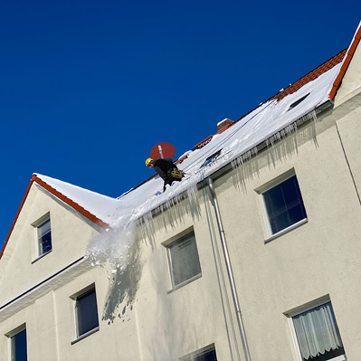 Höhenarbeiter befreit Hausdach im Winter von Schnee. Eiszapfen fallen kontrolliert vom Dach.