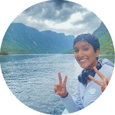 An image of Shivani smiling and making a peace sign on a boat, with a scenic mountain backdrop.