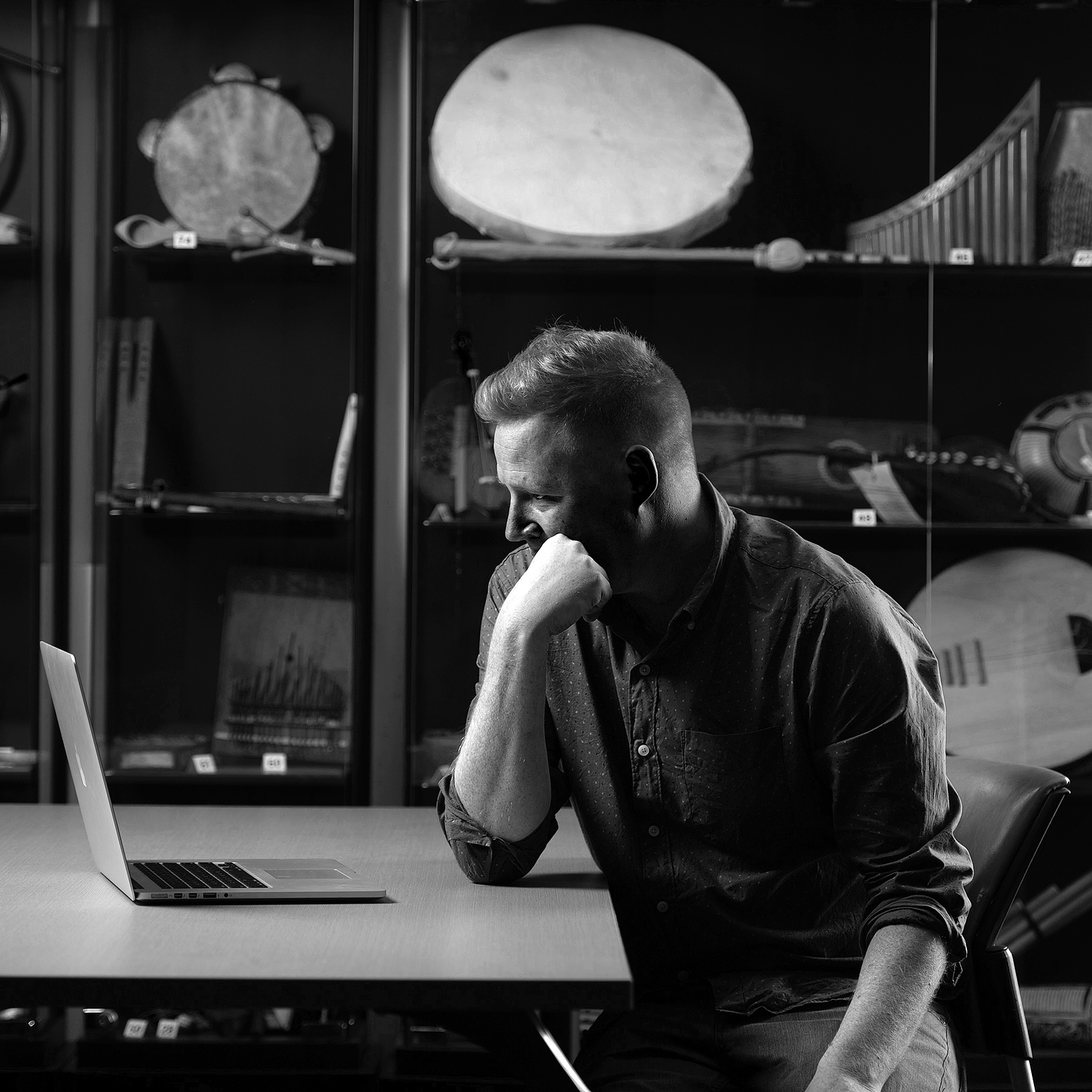 A man sitting at a table in front of a laptop with musical instruments in the background.