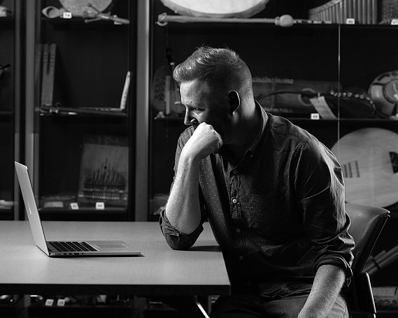 A man sitting at a table in front of a laptop with musical instruments in the background.