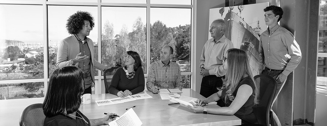 A black and white photo of a medium-sized team discussing brand naming strategy in a conference room that overlooks the Sausalito harbor. Shot at Lexicon Branding in Sausalito, California. 