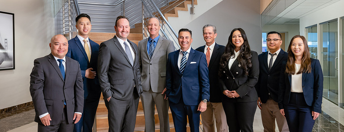 Formal 9-person team/group shot of wealth managers at a First Republic Bank in San Francisco, California. The team members are posed in front of a large staircase in the atrium of their corporate headquarters.