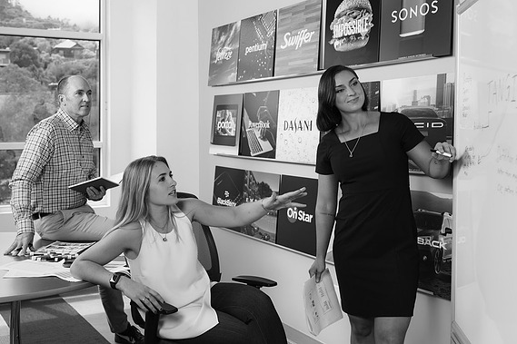 A black and white photo of a team of 3 discussing brand naming strategy next to a white board. Shot at Lexicon Branding in Sausalito, California. 
