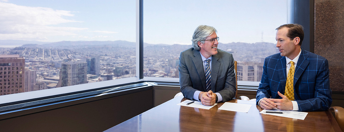 Office lifestyle image of two executives having a meeting on the 40th floor of the 555 California building in the Financial district of San Francisco. Twin Peaks and the rolling city hills of San Francisco are shown through the window behind them.