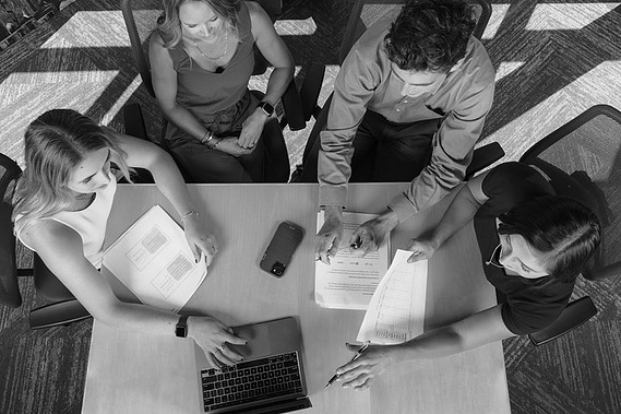 A black and white photo of a team of 4 sitting at a conference table discussing brand naming strategy. The scene is shot from overhead looking down. Shot at Lexicon Branding in Sausalito, California. 