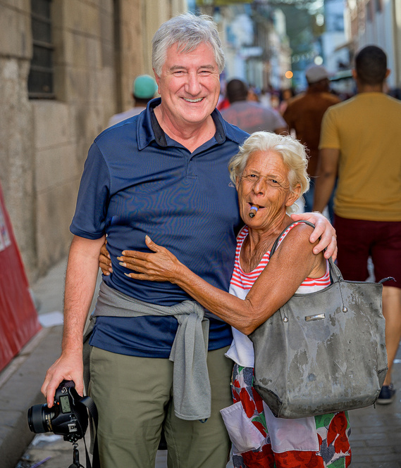 A smiling man with a camera and an elderly woman smoking a cigar pose together in a busy street.
