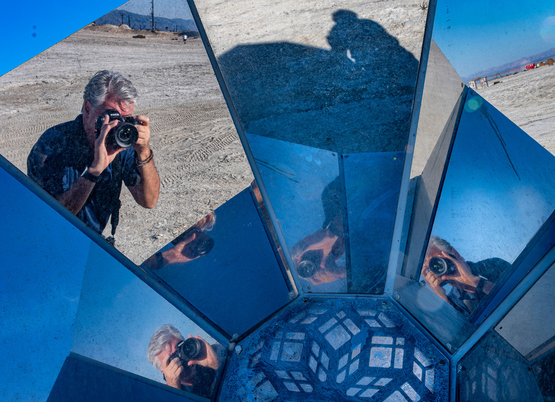 A photographer is reflected multiple times in a mirrored structure, capturing an image in a barren, desert-like landscape.