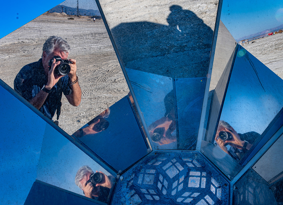 A photographer is reflected multiple times in a mirrored structure, capturing an image in a barren, desert-like landscape.