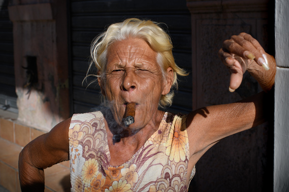 An elderly woman with blonde hair smokes a cigar, wearing a floral dress, and squinting in the sunlight.