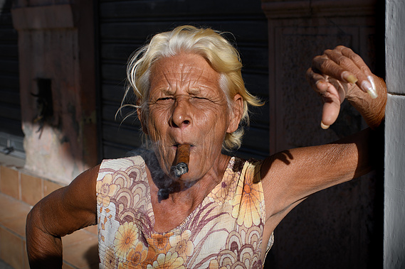 An elderly woman with blonde hair smokes a cigar, wearing a floral dress, and squinting in the sunlight.