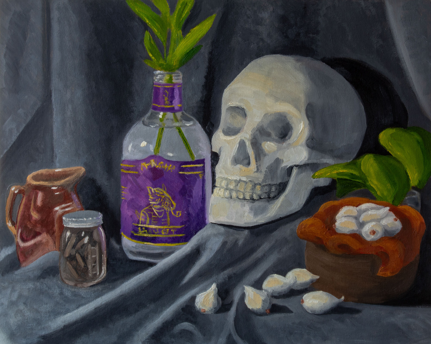 A skull, bowl on garlic cloves, glass jar with plants and other ceramic objects lay on a table covered in fabric.